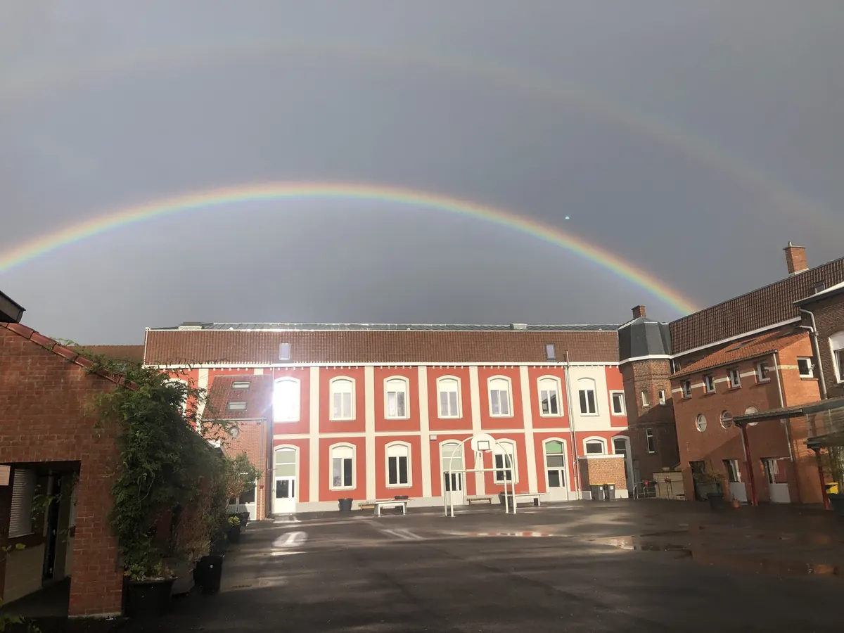 La cour du collège et ses bâtiments en briques sous un double arc-en-ciel.