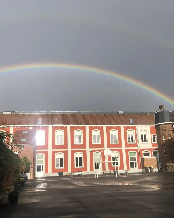 La cour du collège et ses bâtiments en briques sous un double arc-en-ciel.