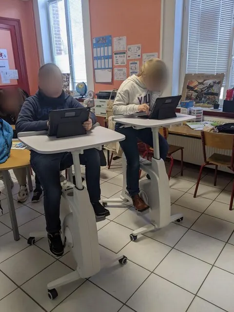 En classe, des élèves travaillent assis sur des vélos de bureau.
